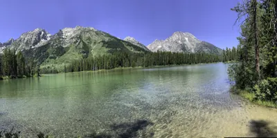 A panoramic view of the clear, shallow waters of a mountain lake reflecting the Teton Range, with Mount Moran prominently visible in Grand Teton National Park, Wyoming.