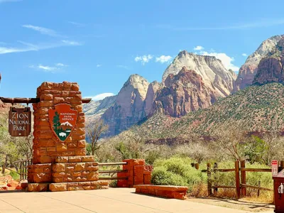 Official stone entrance sign of Zion National Park with the National Park Service logo, backed by towering Navajo sandstone mountains at the South Entrance, Utah.⁠